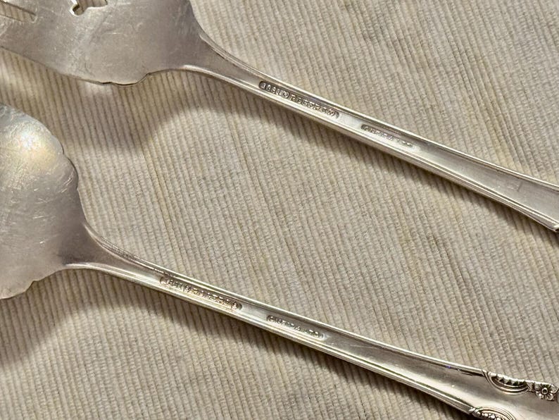 K&ouml;nnte beinhalten: Zwei silberne L&ouml;ffel mit verzierten Griffen. Die L&ouml;ffel tragen den Text "Community Plate" auf der R&uuml;ckseite.