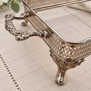 May include: A rectangular silver-toned serving tray featuring an ornate openwork gallery rail, two decorative scrolled handles, and four detailed cabriole-style feet. The metal surface has a polished finish with intricate floral relief patterns.