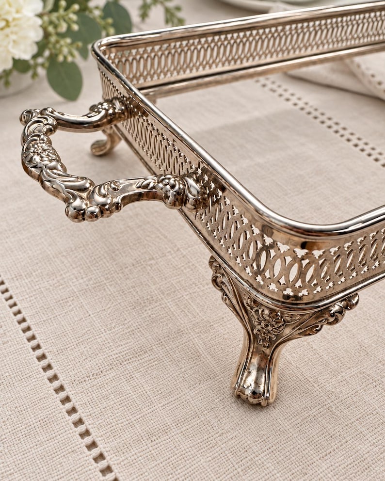 May include: A rectangular silver-toned serving tray featuring an ornate openwork gallery rail, two decorative scrolled handles, and four detailed cabriole-style feet. The metal surface has a polished finish with intricate floral relief patterns.