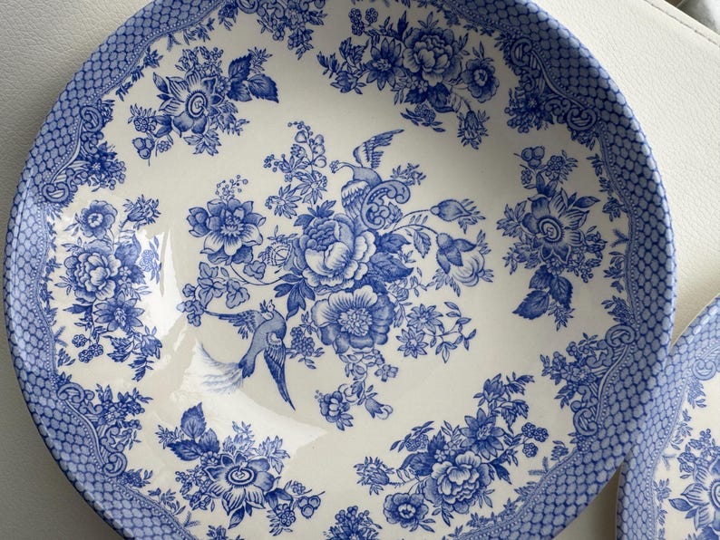 K&ouml;nnte beinhalten: Eine wei&szlig;e Keramiksch&uuml;ssel mit einem blauen und wei&szlig;en Blumenmuster. Die Sch&uuml;ssel zeigt ein sich wiederholendes Muster aus Blumen, Bl&auml;ttern und V&ouml;geln. Die Sch&uuml;ssel ist perfekt zum Servieren von Salaten, Obst oder anderen Gerichten.