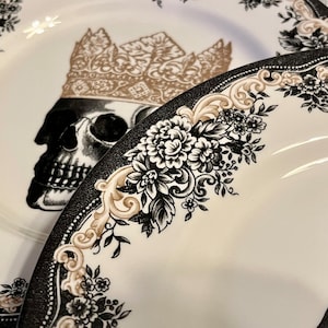 Puede incluir: Dos platos de cer&aacute;mica blanca con una ilustraci&oacute;n de calavera en blanco y negro coronada con una corona dorada. Los bordes est&aacute;n decorados con intrincados patrones florales negros y adornos dorados en forma de volutas.