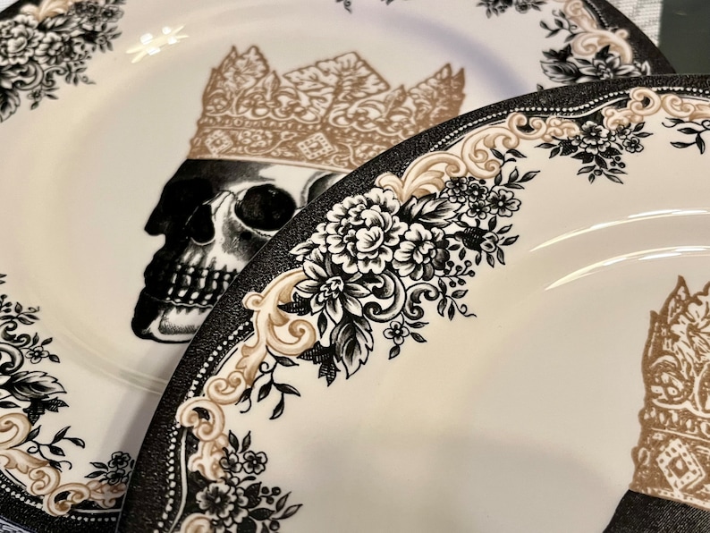 Puede incluir: Dos platos de cer&aacute;mica blanca con una ilustraci&oacute;n de calavera en blanco y negro coronada con una corona dorada. Los bordes est&aacute;n decorados con intrincados patrones florales negros y adornos dorados en forma de volutas.