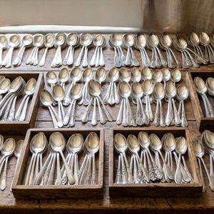 Op de afbeelding: Een assortiment zilveren lepels, uitgestald in houten bakken. De lepels hebben sierlijke handvatten en zijn in nette rijen gerangschikt, wat een verzameling vintage zilverwerk laat zien. De lepels zijn gemaakt van een glanzend, reflecterend metaal.