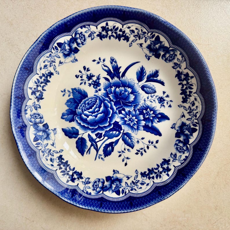 May include: A round ceramic plate with a cream-colored center and a deep blue floral pattern. The design features a central bouquet of roses and leaves, surrounded by a wide, scalloped border decorated with intricate blue floral motifs.
