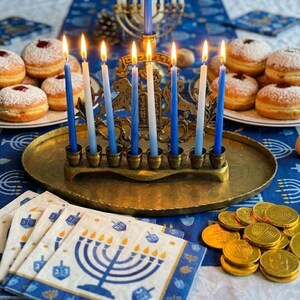 May include: A brass menorah with lit blue and white candles, surrounded by plates of donuts and gold coins. Napkins with menorah designs and a dreidel are also present, creating a festive scene.