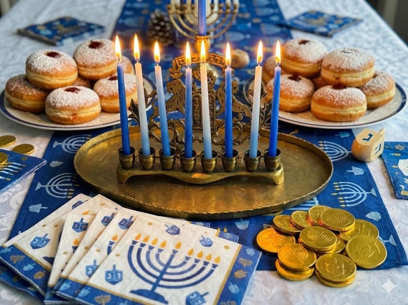 May include: A brass menorah with lit blue and white candles, surrounded by plates of donuts and gold coins. Napkins with menorah designs and a dreidel are also present, creating a festive scene.