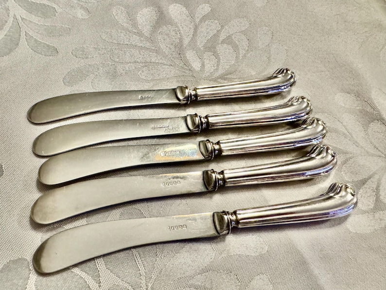 K&ouml;nnte beinhalten: F&uuml;nf silberne Buttermesser mit gebogenen Griffen und abgerundeten Klingen sind auf einer gemusterten Oberfl&auml;che angeordnet. Jedes Messer hat einen dekorativen Griff und eine leicht gebogene Klinge. Die Messer bestehen aus gl&auml;nzendem, reflektierendem Metall.