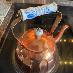 May include: A copper tea kettle with a fluted body and a curved spout. The handle is made of white ceramic with blue floral patterns and brass-toned metal supports. A matching white ceramic knob with a blue floral design sits on the lid.