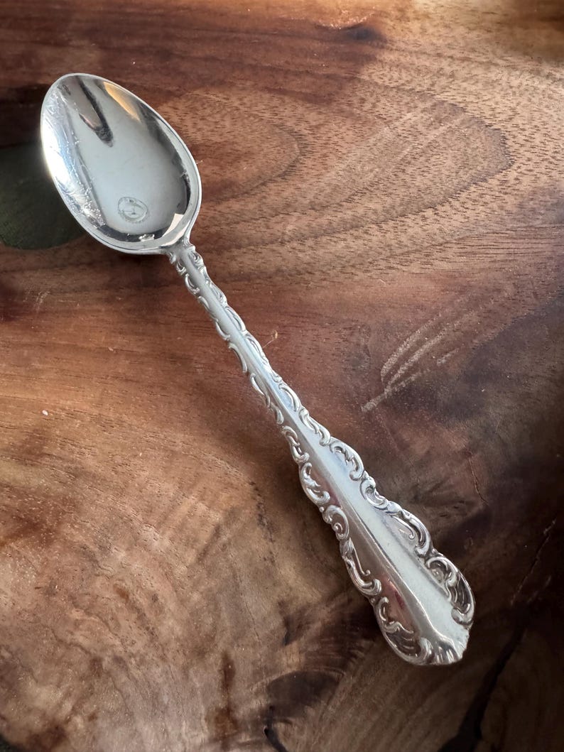 May include: A silver-colored metal spoon featuring an oval bowl and a long handle decorated with intricate embossed scrollwork and floral patterns along the edges, terminating in a flared, rounded end with a polished finish.