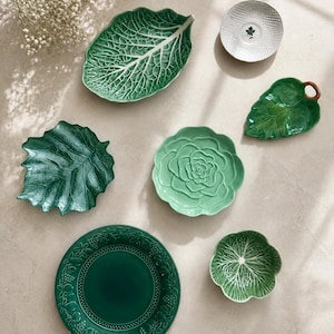 Puede incluir: Una colecci&oacute;n de vajilla de cer&aacute;mica que incluye una fuente grande con forma de hoja verde, un platillo blanco texturizado, un plato con forma de hoja verde con tallo marr&oacute;n, un plato verde oscuro con bordes de frutas y dos cuencos verdes peque&ntilde;os.
