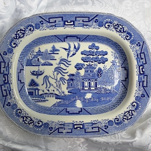 May include: An oval ceramic platter featuring a blue and white willow pattern. The central design depicts a landscape with a pagoda, a bridge, a willow tree, and two birds in flight. The wide rim is decorated with a geometric and floral border.