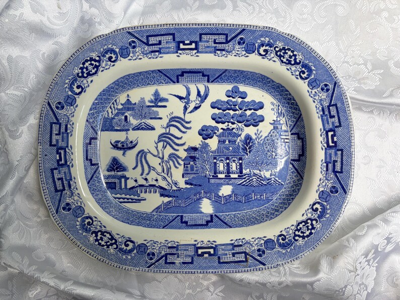 May include: An oval ceramic platter featuring a blue and white willow pattern. The central design depicts a landscape with a pagoda, a bridge, a willow tree, and two birds in flight. The wide rim is decorated with a geometric and floral border.