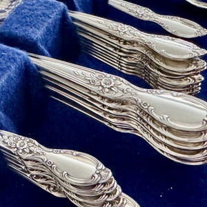 K&ouml;nnte beinhalten: Ein Stapel versilbertes Besteck mit verzierten Blumenmustern. Das Besteck ist in einem blauen Samt-Etui angeordnet.