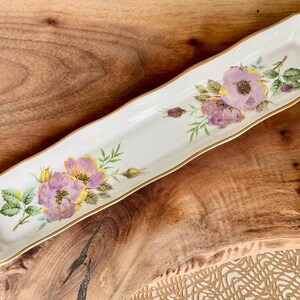 May include: A white ceramic serving dish with a gold rim and a floral design featuring purple and yellow roses.