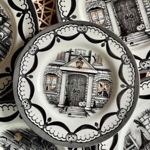 K&ouml;nnte beinhalten: Ein Set aus vier schwarzen und wei&szlig;en Halloween-Keramiktellern. Jeder Teller zeigt ein Spukhaus mit einer schwarzen T&uuml;r und einer schwarzen Katze. Die Teller sind mit einem schwarzen und wei&szlig;en gewellten Rand und einem schwarzen und wei&szlig;en Bandmuster verziert. Die Teller sind perfekt f&uuml;r eine Halloween-Dinnerparty.