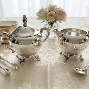 Puede incluir: Un azucarero de plata pulida con tapa y remate, una jarra de leche a juego, unas pinzas para az&uacute;car de plata y una peque&ntilde;a cuchara de plata. Cada pieza presenta pies y asas curvados y ornamentados con un acabado met&aacute;lico brillante.