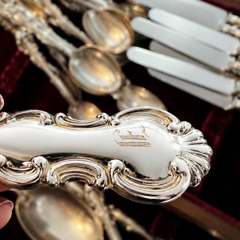 Puede incluir: Primer plano de cucharas y cuchillos de plata ornamentados. Las cucharas tienen un dise&ntilde;o de mango detallado con un monograma. La cuberter&iacute;a est&aacute; dispuesta sobre una superficie de terciopelo rojo oscuro, lo que sugiere una colecci&oacute;n vintage o antigua.