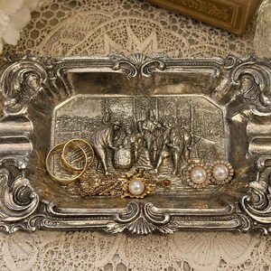 May include: A rectangular silver-toned metal tray with ornate, scrolled edges and a central embossed relief scene depicting figures gathered around a barrel. The tray contains two gold-toned rings, a gold chain, and two pearl-accented earrings.