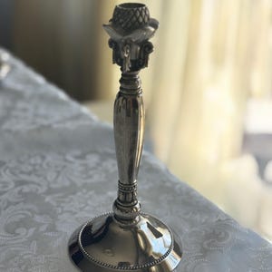 May include: A silver-toned metal candlestick holder featuring a smooth, tapered central stem, a wide circular base with a beaded edge, and a decorative top section with a textured, pineapple-like pattern and ornate scrollwork details.