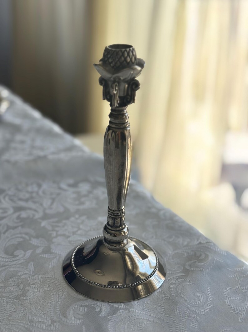 May include: A silver-toned metal candlestick holder featuring a smooth, tapered central stem, a wide circular base with a beaded edge, and a decorative top section with a textured, pineapple-like pattern and ornate scrollwork details.