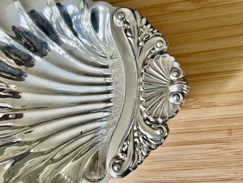 May include: A silver serving dish shaped like a seashell with ornate details. The dish is highly polished and reflects light.