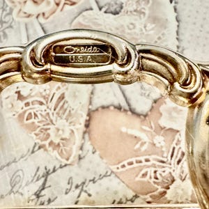 May include: A silver-toned metal handle featuring an ornate, curved design with a raised rectangular plaque stamped with the text Oneida U.S.A. The handle is attached to a metallic base with a decorative, perforated edge pattern along the bottom.