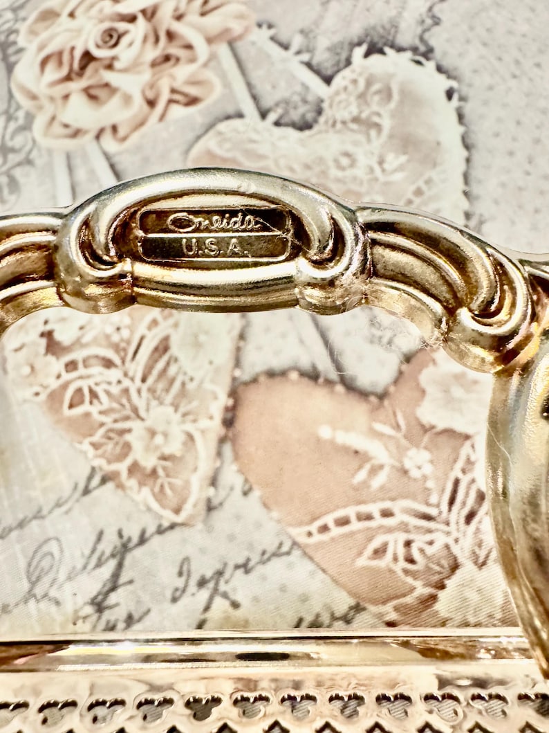 May include: A silver-toned metal handle featuring an ornate, curved design with a raised rectangular plaque stamped with the text Oneida U.S.A. The handle is attached to a metallic base with a decorative, perforated edge pattern along the bottom.