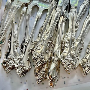 May include: Close-up of a collection of ornate silver-colored flatware, including forks, knives, and spoons. The handles feature a detailed floral and scroll design. The silverware is arranged on a white surface with colorful polka dots.