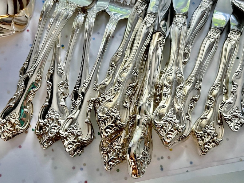 May include: Close-up of a collection of ornate silver-colored flatware, including forks, knives, and spoons. The handles feature a detailed floral and scroll design. The silverware is arranged on a white surface with colorful polka dots.