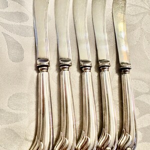 K&ouml;nnte beinhalten: F&uuml;nf silberne Buttermesser mit gebogenen Griffen und abgerundeten Klingen. Die Griffe haben ein dekoratives, wirbelndes Design an der Basis. Die Messer sind in einer Reihe angeordnet und zeigen ihre polierten, reflektierenden Oberfl&auml;chen.