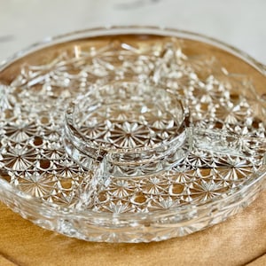 May include: Clear glass serving dish with a starburst pattern. The dish is divided into sections, with a central circular compartment and three surrounding sections. The dish sits on a wooden tray.