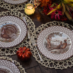 May include: Four brown and white patterned dinner plates with a woven placemat underneath each. The plates feature a turkey and pheasant design. A small orange pumpkin sits on the table.