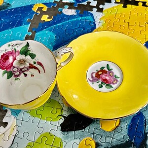 Pu&ograve; includere: Un set di tazza da t&egrave; e piattino in porcellana gialla con interno bianco decorato da un bouquet di fiori rosa, bianchi e viola. La tazza presenta un bordo smerlato con finiture dorate e un manico decorativo, con un motivo floreale sul piattino.