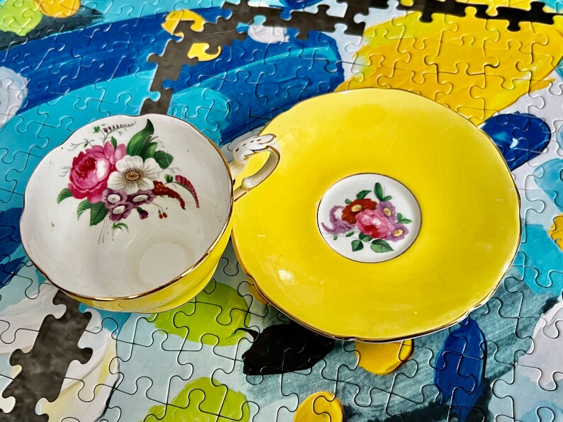 Pu&ograve; includere: Un set di tazza da t&egrave; e piattino in porcellana gialla con interno bianco decorato da un bouquet di fiori rosa, bianchi e viola. La tazza presenta un bordo smerlato con finiture dorate e un manico decorativo, con un motivo floreale sul piattino.
