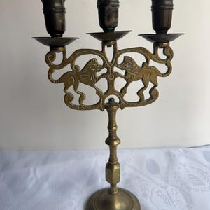 Pu&ograve; includere: Un candelabro in ottone con tre portacandele. Il pezzo decorativo presenta due figure di leone sul davanti. La base &egrave; rotonda e l'altezza totale &egrave; di circa 25 cm.