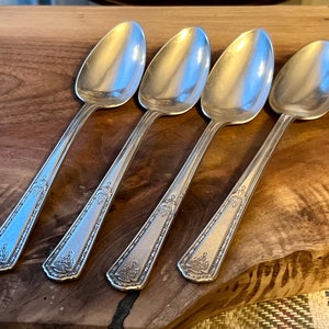 Pu&ograve; includere: Quattro cucchiai d'argento con manici decorati sono disposti su una tavola di legno. I cucchiai hanno un design classico con una coppa arrotondata e un motivo decorativo sul manico. I cucchiai sono riflettenti.