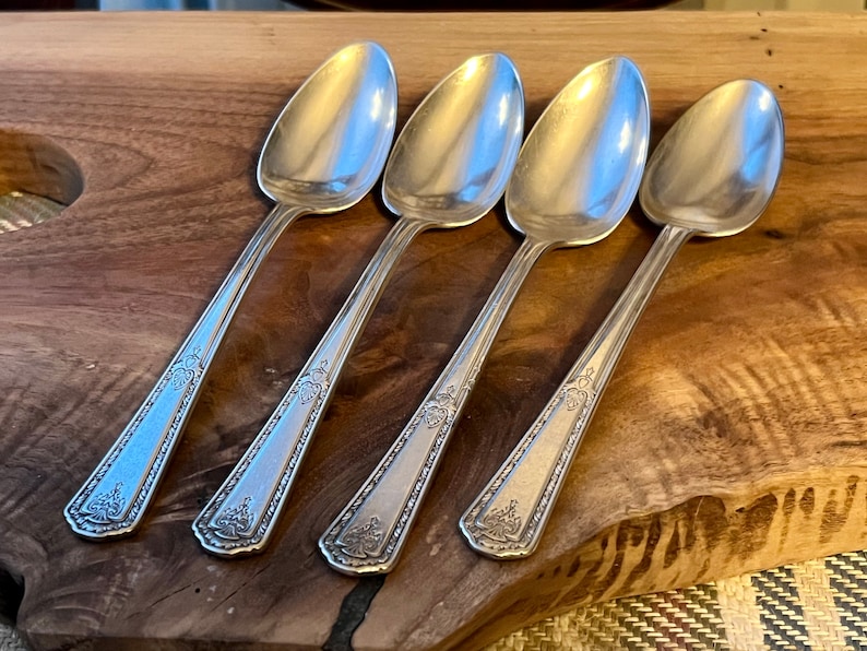 Pu&ograve; includere: Quattro cucchiai d'argento con manici decorati sono disposti su una tavola di legno. I cucchiai hanno un design classico con una coppa arrotondata e un motivo decorativo sul manico. I cucchiai sono riflettenti.