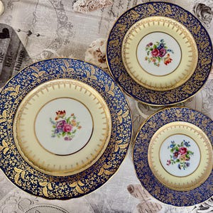 May include: Three antique porcelain plates with a floral design. The plates have a cream-colored center with a bouquet of flowers and a navy blue border with gold leaf detailing. The plates are of different sizes.