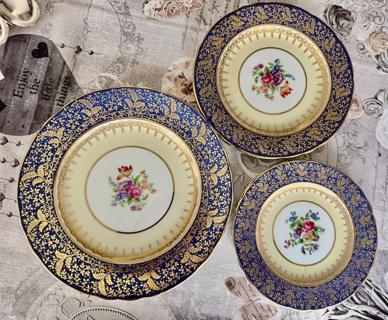 May include: Three antique porcelain plates with a floral design. The plates have a cream-colored center with a bouquet of flowers and a navy blue border with gold leaf detailing. The plates are of different sizes.
