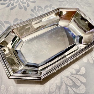 K&ouml;nnte beinhalten: Ein poliertes, silberfarbenes Metalltablett mit achteckiger Form und einem mehrstufigen Rand. Das Tablett verf&uuml;gt &uuml;ber eine glatte, reflektierende Oberfl&auml;che mit abgewinkelten Seiten, die ein geometrisches, facettiertes Erscheinungsbild erzeugen.