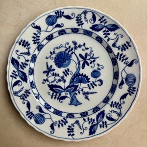 May include: A round white ceramic plate with a scalloped edge, featuring a blue underglaze pattern of stylized floral motifs, vines, and pomegranate-like shapes. The central design depicts a large blossom, while the rim displays repeating botanical elements.