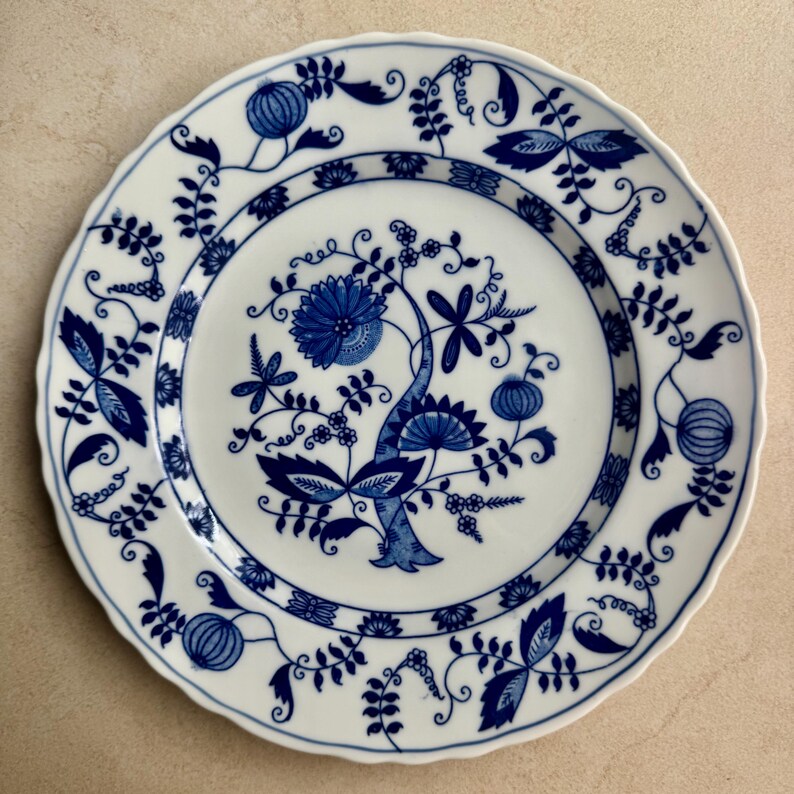 May include: A round white ceramic plate with a scalloped edge, featuring a blue underglaze pattern of stylized floral motifs, vines, and pomegranate-like shapes. The central design depicts a large blossom, while the rim displays repeating botanical elements.