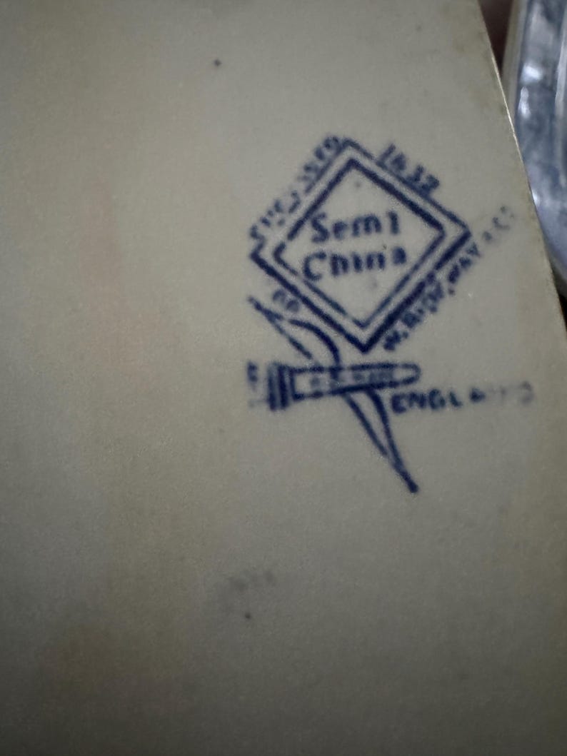 May include: A cream-colored ceramic surface featuring a dark blue stamped maker's mark. The mark includes a diamond shape with the text Semi China, the number 1632, the name W. Ridgway & Co, and the word England, accompanied by a small graphic icon.