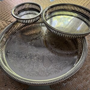 May include: A set of three silver-toned metal serving pieces including one large round tray with an ornate engraved center and two smaller round bowls. Each piece features a raised gallery rim with a vertical slotted design and a twisted rope edge.