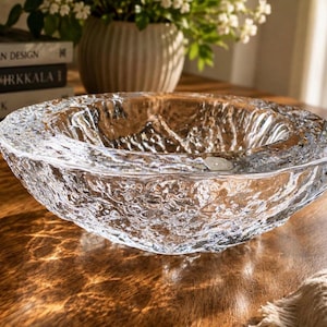 May include: A clear glass bowl featuring a thick, irregular, and textured exterior surface that mimics the appearance of melting ice or flowing water. The bowl has a wide, smooth, and rounded rim with a deep, concave center and a heavy base.