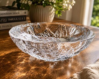 May include: A clear glass bowl featuring a thick, irregular, and textured exterior surface that mimics the appearance of melting ice or flowing water. The bowl has a wide, smooth, and rounded rim with a deep, concave centre and a heavy base.
