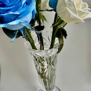 May include: A clear glass vase with a starburst and vertical cut pattern containing a bouquet of fresh roses. The arrangement features two large vibrant blue roses and two white roses with green stems and leaves visible inside the glass.