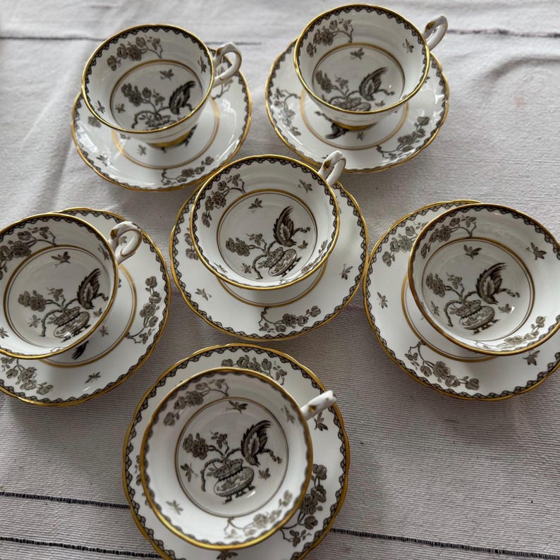 May include: Set of six teacups and saucers with a black and white floral design. The cups and saucers have a gold trim.