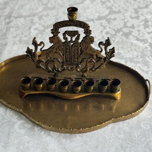 May include: A brass menorah with a decorative design featuring lions and Hebrew text, set on an oval tray. The menorah has eight candle holders and a central holder for the shamash candle.