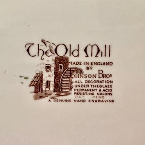 Op de afbeelding: Een bruin keramisch fabrieksmerk met een illustratie van een watermolen en de tekst: The Old Mill, Made in England by Johnson Bros, All decoration under the glaze permanent & acid resisting colors, Pat Pend, A genuine hand engraving.